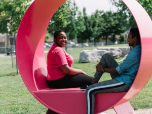 Photo of two female students sitting in an artistic red bench in a park