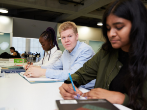 Three students studying at a desk. One student looking straight into the camera.